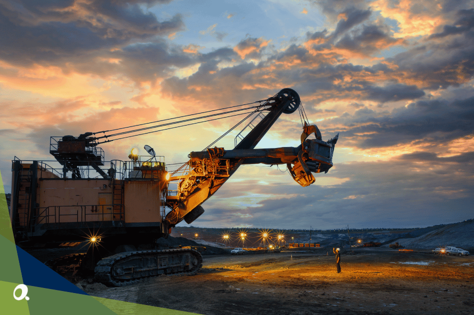 Large excavator operating at a construction or mining site during sunset, with a worker standing nearby.