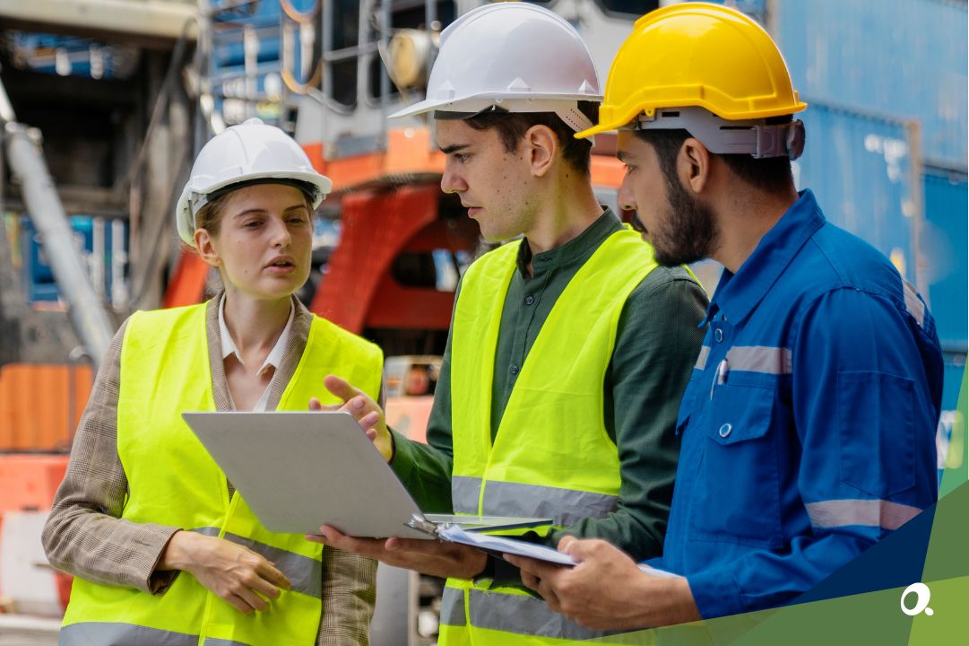 Three logistics workers in safety gear reviewing shipping data on site, discussing supply chain performance.