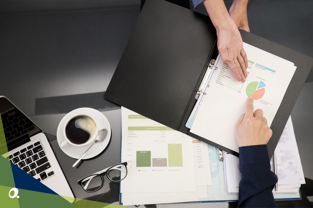 Two people reviewing financial documents with charts in a binder, alongside a laptop, glasses and a cup of coffee on the table.