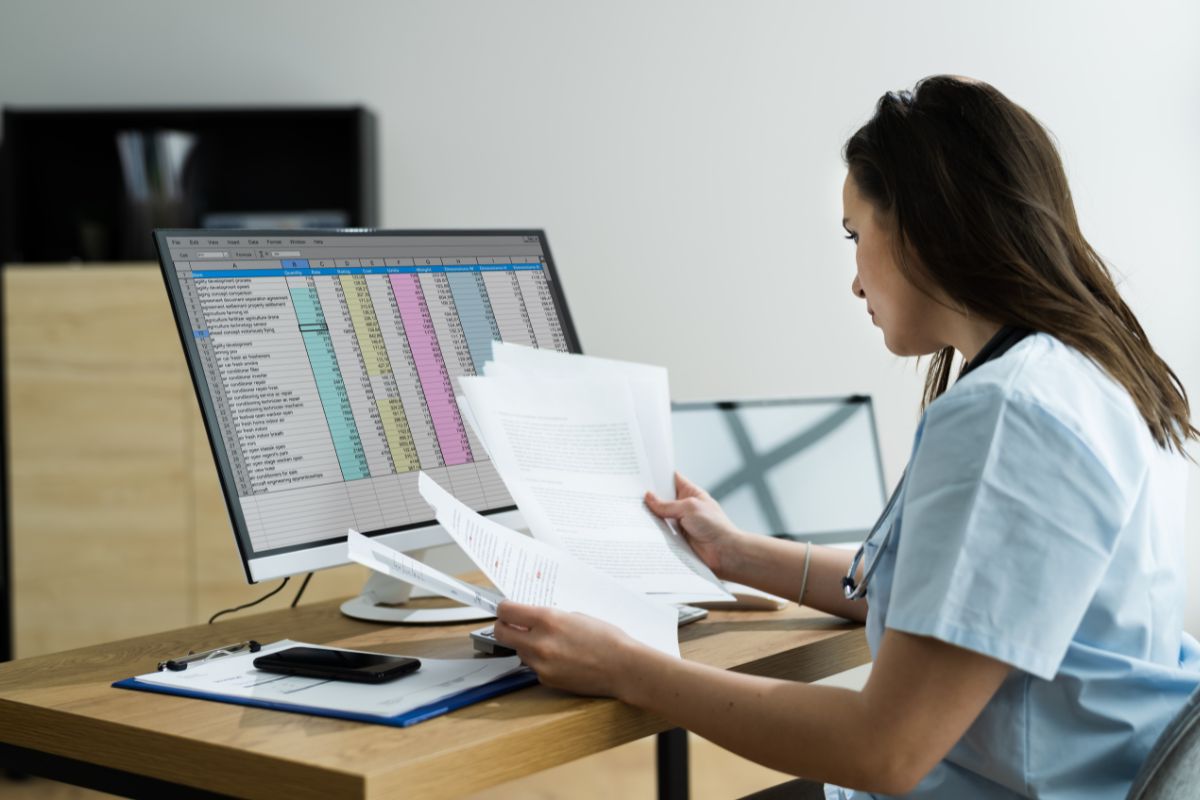 Woman holding a bunch of papers in front of a computer with a spreadsheet program